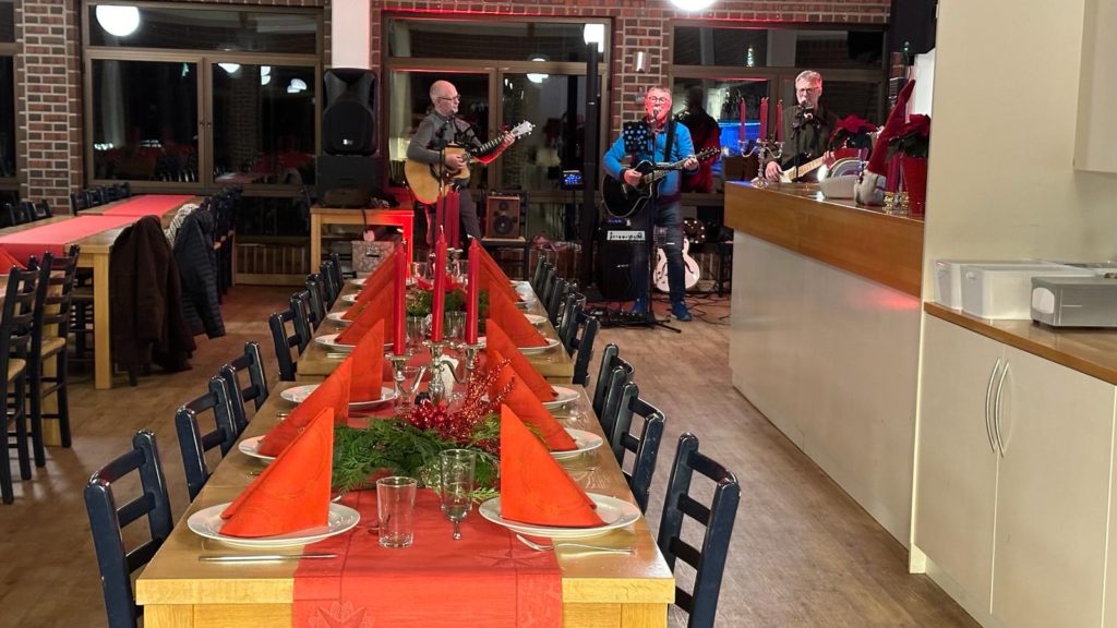 Bord som er dekket klart for julebordet på Østre Bolæren i 2023, i bakgrunnen kan man se tre menn som spiller på gitar og synger. Foto.